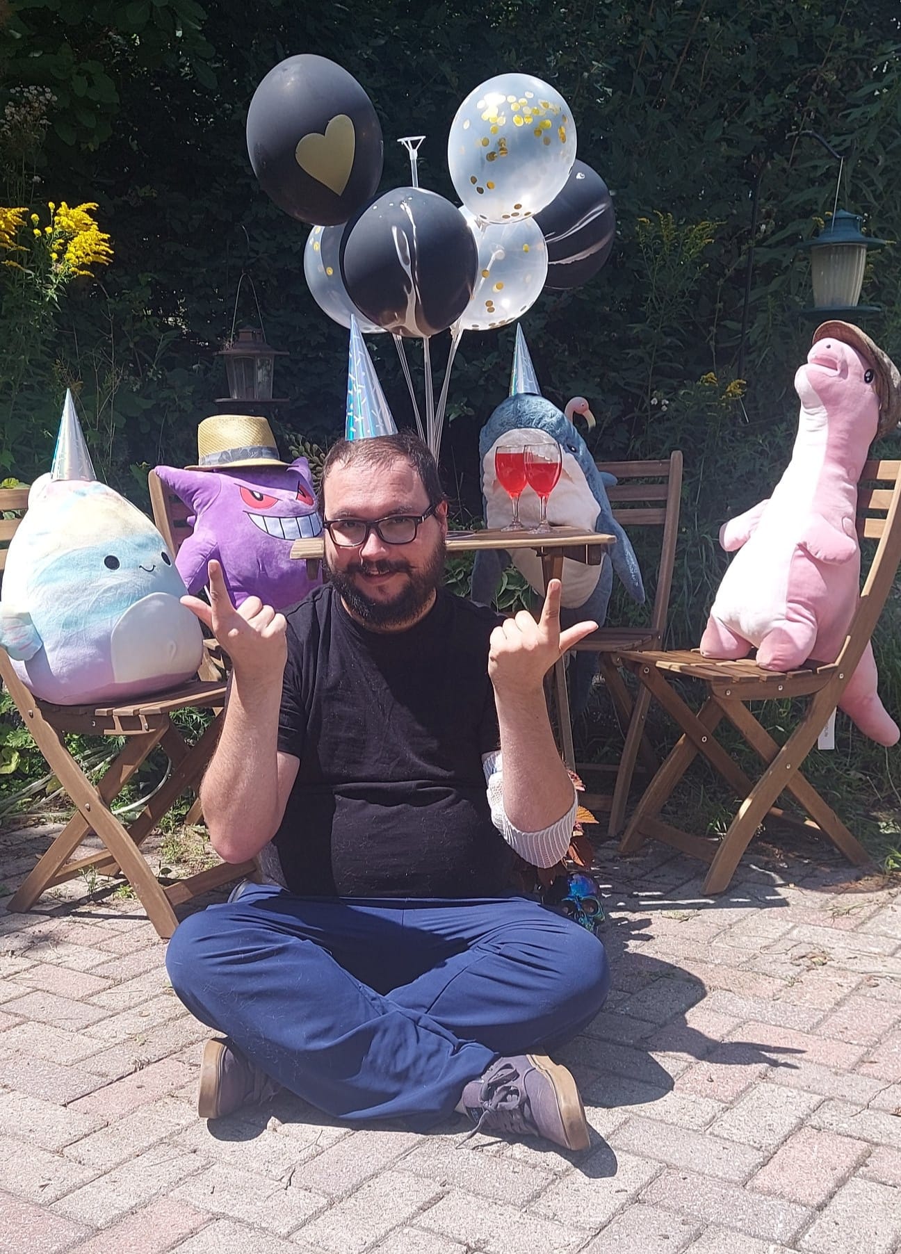 A man with facial hair and a felt sun hat sits on the ground in front of a patio set hosting a stuffed animal patio party, Photo 5